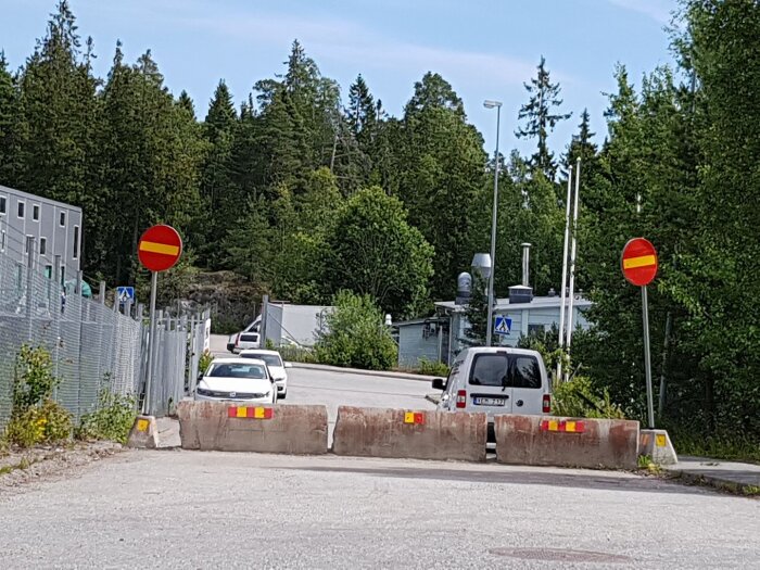 Väg med betongblock och förbjuden infart-skyltar, två bilar syns i bakgrunden vid en skogskant och byggnad.
