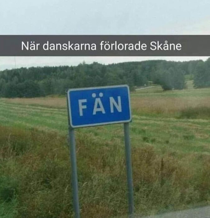 Vägmärke med texten "FÄN" i landsbygdsmiljö med skogar och fält i bakgrunden.