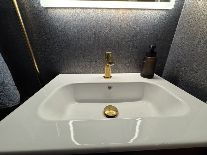 Modern handfat med guldfärgad kran och mörk strukturtapet på vägg, svart tvålpump placerad bredvid handfatet.
