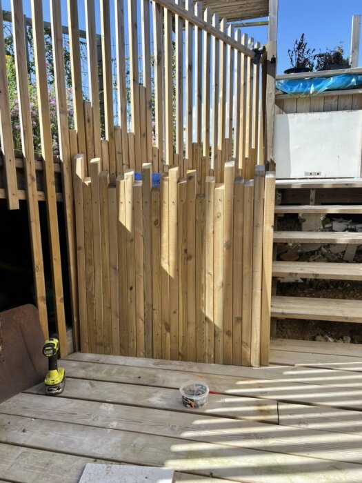 Nybyggd trägrind och staket i källartrappa med en skruvdragare och en burk skruvar på trädäcket i solljus.