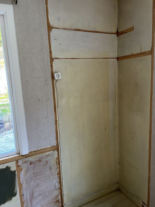 Hörn av rum med vävtapet delvis borttagen, synliga släta och ojämna ytor samt eluttag på vägg och fönster till vänster.