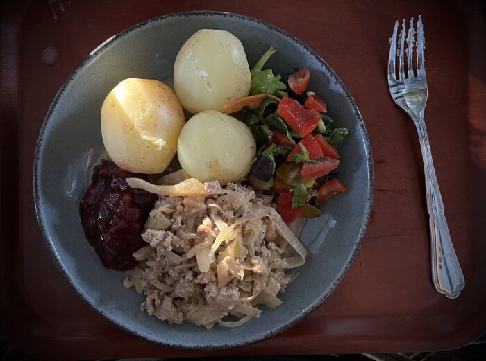Tallrik med kålmaja, kokt potatis, lingonsylt och sallad, serverad på en bricka med gaffel.