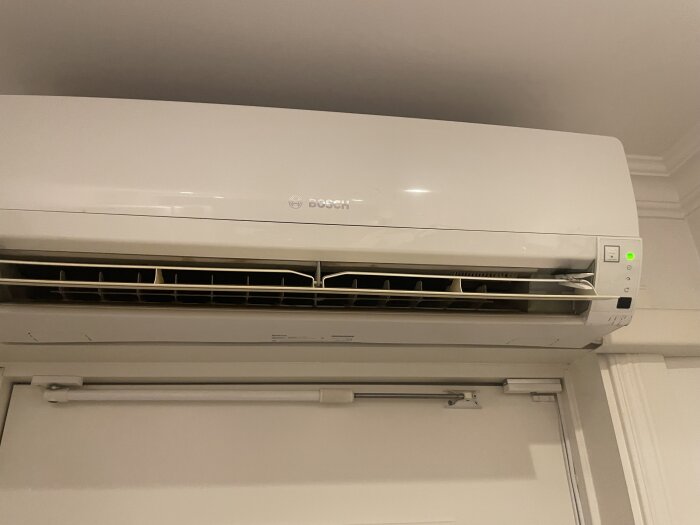 Vit Bosch luftvärmepump inomhusenhet med stillastående vingar och en grön indikatorlampa, monterad ovanför en dörr.