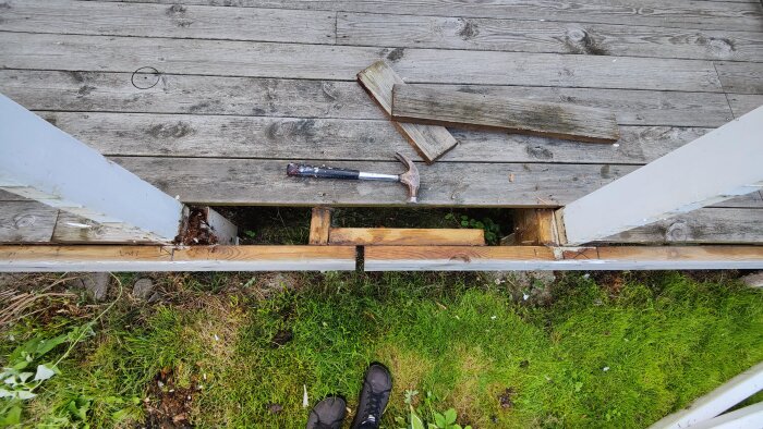 Skadad altan med borttagen bärlina, två stolpar och en hammare synliga. Gräs under altanen.