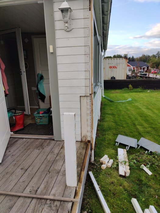 Altan med vit panel, trasig knutbräda och löst bråte på gräset framför. Öppen dörr med röda stövlar inomhus.