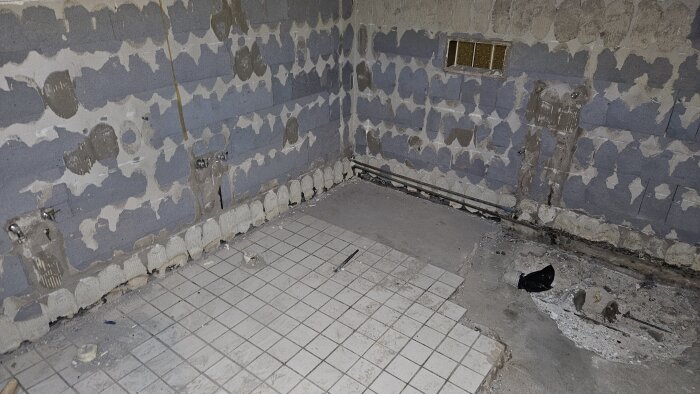 Renoverat badrum med delvis avlägsnat kakel, blottlagda lättbetongväggar och en del kakelplattor kvar på golvet. Verktyg och skräp ligger kvar.