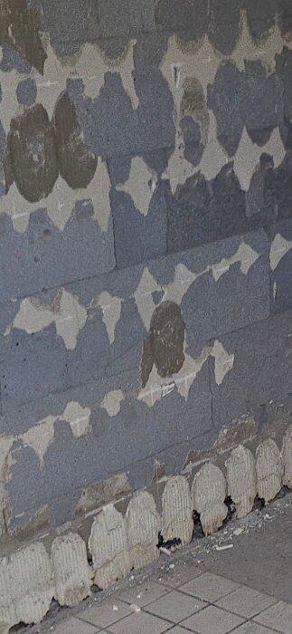 Vägg av lättbetong synlig efter att kaklet tagits bort, med rester av gammalt kakelfix kvar. Golvet är täckt av fyrkantiga plattor.