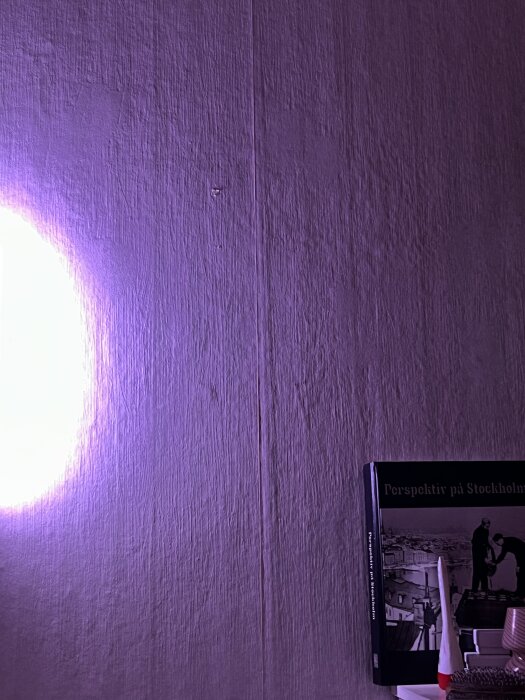 Strukturerad vägg med synliga luftfickor och bok på hylla, belyst av lila sken, möjligen en strukturtapet från 50-talet.