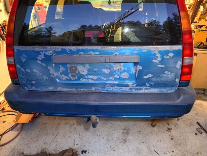 Bakluckan på en blå bil med borttagna emblem och rostfläckar, redo för slipning och grundning. Verktyg och sladdar synliga i bakgrunden.