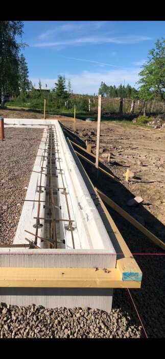 Byggplats med formsättning för grundläggning, grus och armeringsjärn synliga, omgiven av skog och mark preparerad för gjutning.