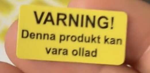 Gul varningsetikett med texten "VARNING! Denna produkt kan vara ollad," hålls i en hand.