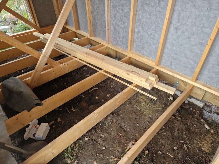 Byggkonstruktion av en komplementbyggnad med träbalkar och jordgolv; material och verktyg syns i bilden.