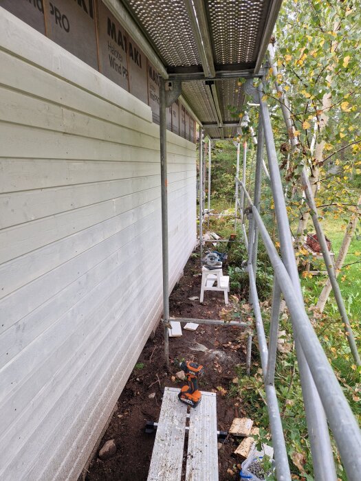 Byggnadsställning vid en fasad av nybyggt träbeklätt hus i skogsmiljö, med verktyg och material längs marken.