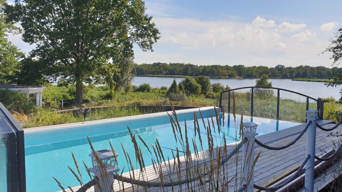 Infinitypool med trädäck och glasräcke, omgiven av grönska, med utsikt över havet i soligt väder.