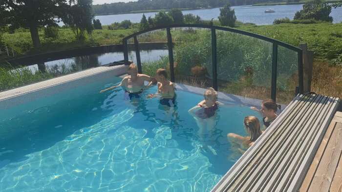Flera personer badar i en pool med en infinity-kant mot en sjö. Poolens design inkluderar glasskydd och en trädäckad omgivning.