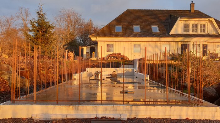 Betongplatta för poolkonstruktion med armeringsjärn och termoblock. Ett hus i bakgrunden. Solens ljus ger en varm ton över platsen.