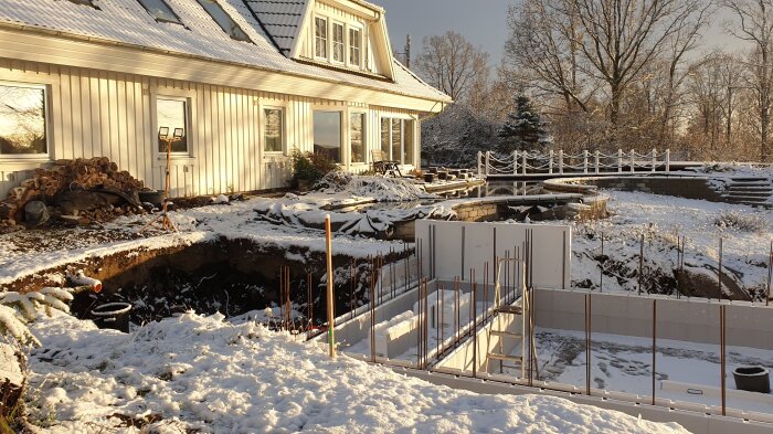 Snötäckt byggplats med en delvis färdigställd poolkonstruktion av termoblock framför ett hus i vintersol.