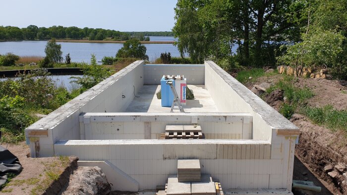 Byggarbetsplats, rektangulär poolkonstruktion med fyllda thermoblock, omgiven av natur och vatten i bakgrunden.