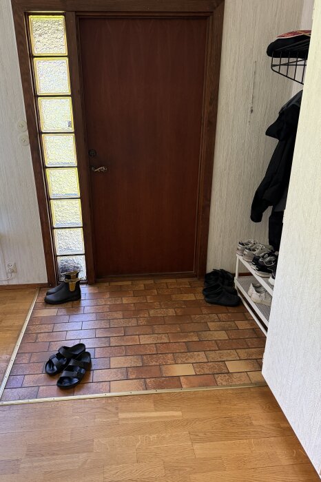 Entré med bruna klinkerplattor, trätröskel och glasblock till vänster om en brun dörr, skoställ och klädhängare till höger.