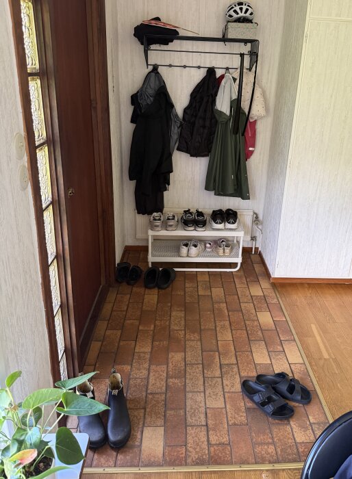 Entré med brun klinkergolv, klädhängare på väggen, skoställ med skor, samt en växt i förgrunden.
