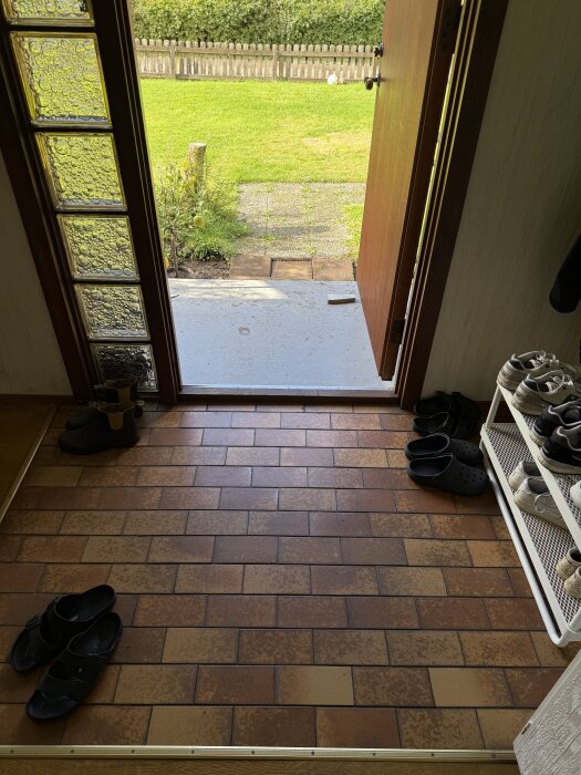 Entré med brunt klinkergolv, öppen ytterdörr och vy mot gräsmatta utomhus; skor vid dörren och skohylla syns.