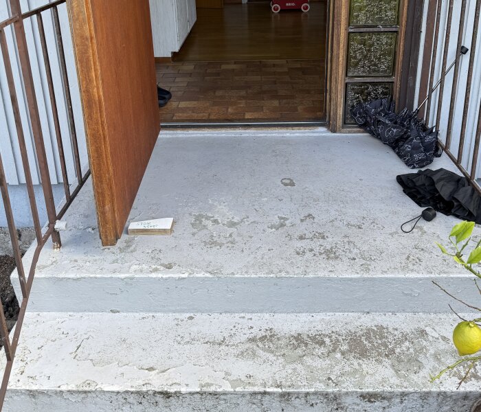 Öppen ytterdörr med trappa och entrégolv, föreslås sänkas för att skapa en genkan. Kläder och paraply bredvid dörren, synliga insidan golvplattor.