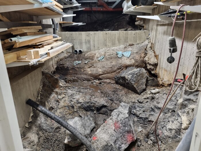 Bilden visar ett inomhusutrymme med synligt berg, två hyllor med diverse verktyg och virke, samt några rör och kablar i förgrunden.