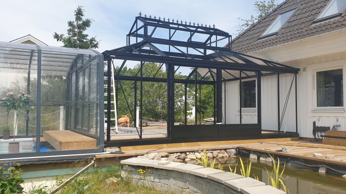 Modernt växthus i svart stål och glas bredvid en pool, anslutet till ett hus, med landskapsträdgård och byggutrustning synlig.
