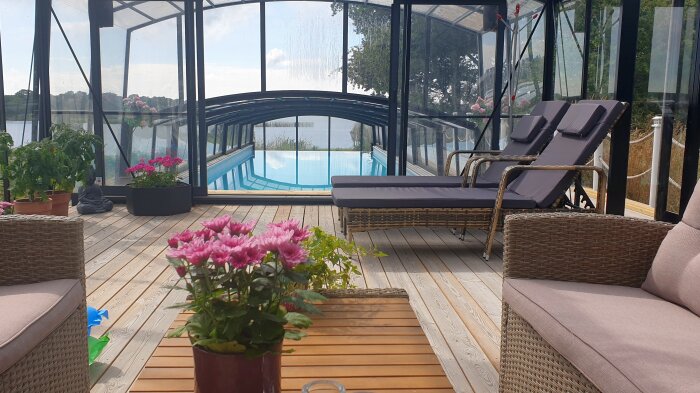Växthus vid en pool med trätrall, loungestolar och blomkrukor, beläget nära vatten och omgivet av gröna växter.