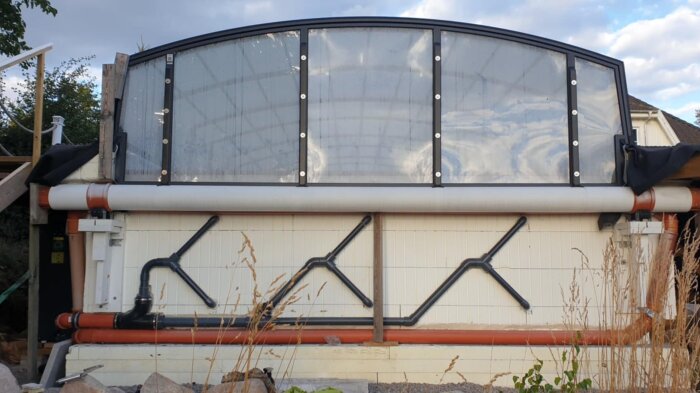 Öppningsbart gavelfönster i pooltak med synliga rör och växter i förgrunden, himmel syns i bakgrunden.