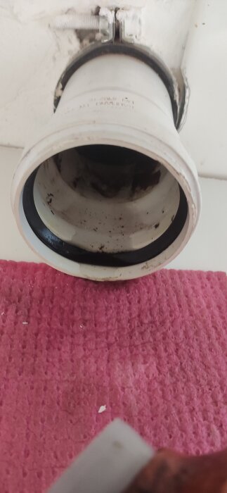 Närbild av avloppsrörsanslutning med svart tätningsring och smuts, omgivet av vitt material, över en rosa trasa.