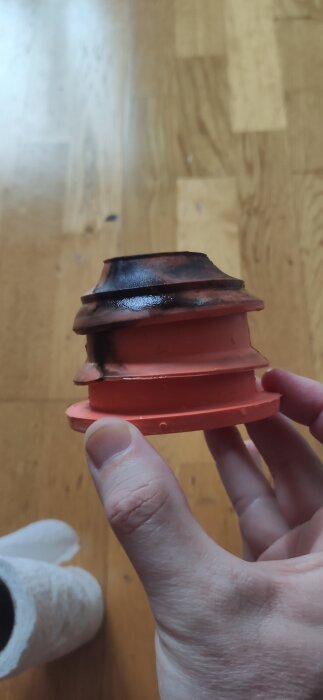 Hand håller i en orange avloppsdamask med svart gummitätning, synlig slitage. Texturerad golvyta i bakgrunden.