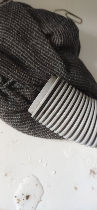 En avloppsslang och en blöt pöl på ett vitt golv visas. Slangen ligger ovanpå en grå trasa.