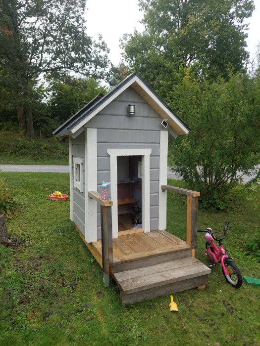 Grå lekstuga med vitt foder, träveranda och liten rosa cykel bredvid, omgiven av träd och gräsmatta.