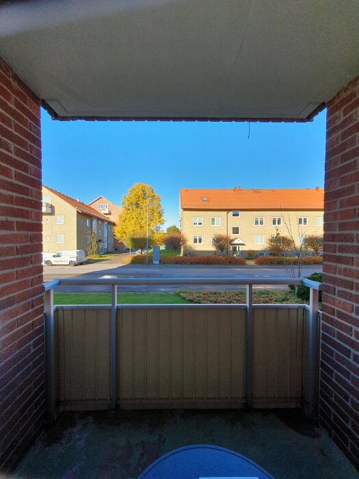 Balkong med tegelväggar och utsikt mot gatan, omgiven av flerfamiljshus och träd under klarblå himmel.