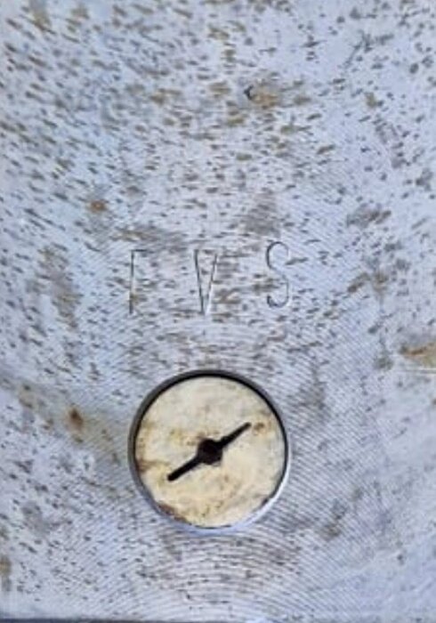 Liten lucka märkt "FVS" på metallplåt, cirkulär skruv eller vridmekanism i centrum.