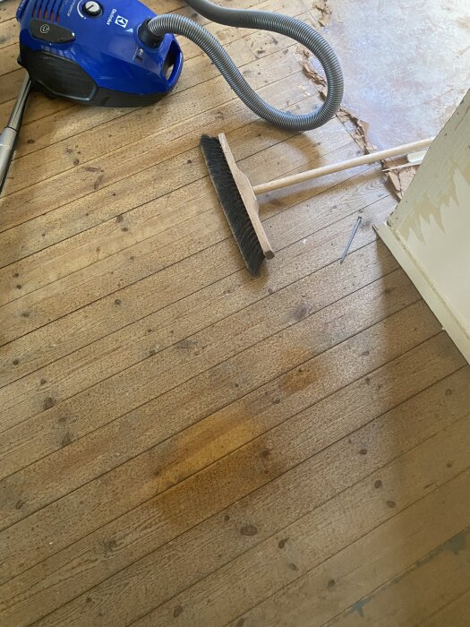 Trägolv från 1930 med synliga prickar, en sopborste och en dammsugare; rester av upplöst vinylgolv vid kanterna.