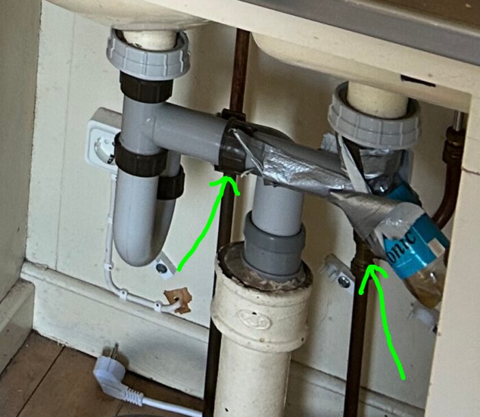 Avlopp under diskbänk med två gröna pilar som pekar på rörkopplingar; silvertape och plast används runt kopplingarna.
