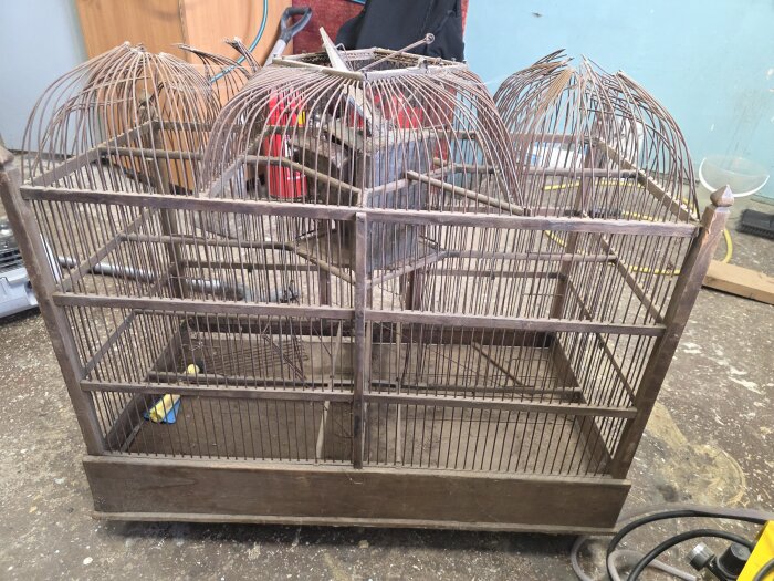 Antik fågelbur av trä och metall, troligtvis från sent 1800-tal till början av 1900-talet, står i ett verkstadsutrymme.