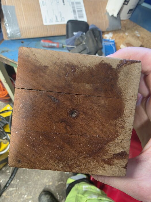 Person håller mörkt trästycke från fågelbur, visar slitage och färgvariationer, möjligtvis påverkad av bivax. Bakgrund visar verktyg och arbetsbänk.