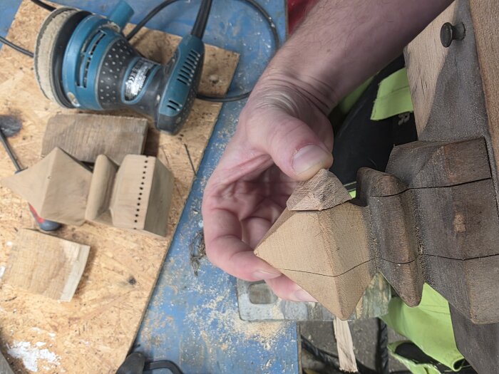 Hand håller trädetalj från fågelbur, slipmaskin och trädelar synliga i bakgrunden.