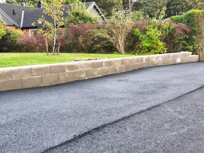 Nyasfalterad kommunal väg med ojämn yta bredvid mur av betongblock, omgiven av grönska och buskar. Visar tydlig nivåskillnad i asfalten.