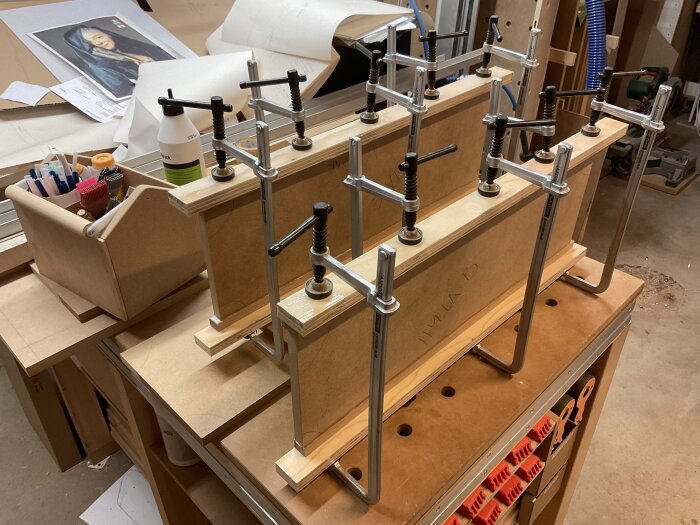 Verkstad med mdf-paneler fastsatta med klämmor, verktyg och pennor i en låda, samt en bild av en målning i bakgrunden.