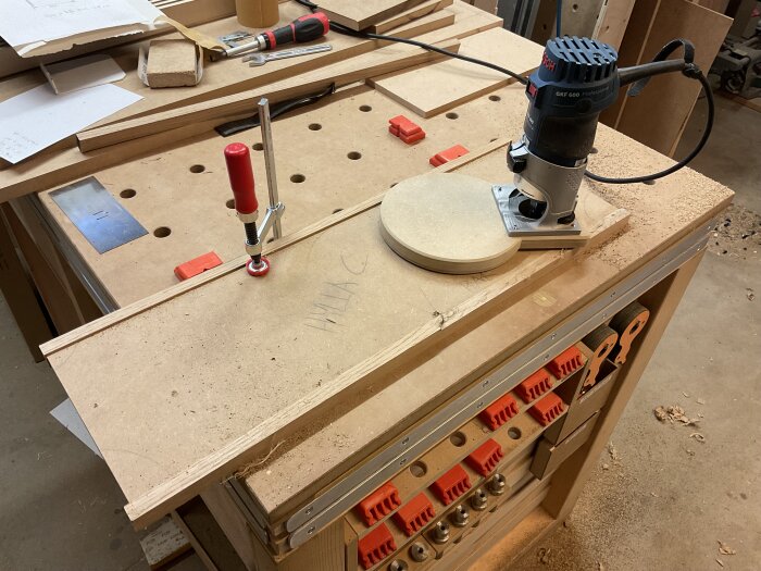 Arbetsyta med trärundel och fräs i ett snickeriprojekt, med diverse verktyg och material i bakgrunden.