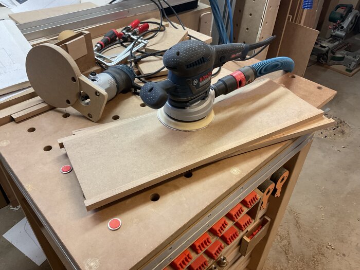 Rundslipmaskin bearbetar MDF-skiva på arbetsbänk i verkstad med verktyg och material synliga i bakgrunden.