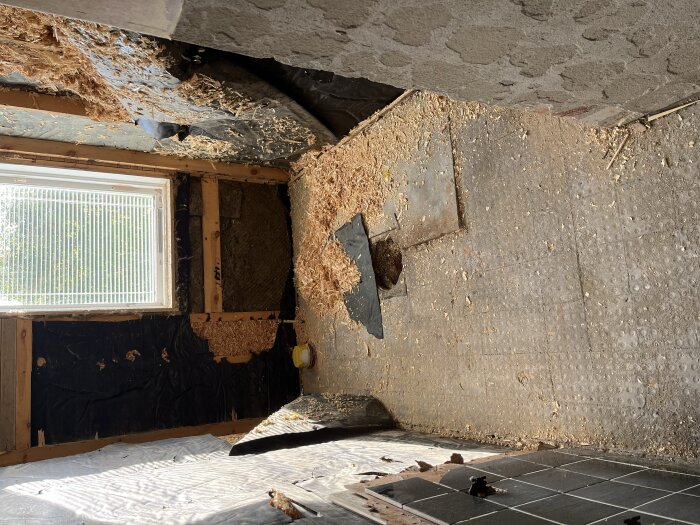Badrum med riven vägg och golv, blottad isolering, betonggolv och sågspån; förekomst av vattenskada ses under fönster med persienner.