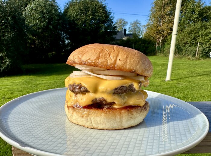 Dubbel cheeseburgare med ost, silverlök och lingon-bbqsås på en tallrik, med grön trädgård i bakgrunden.