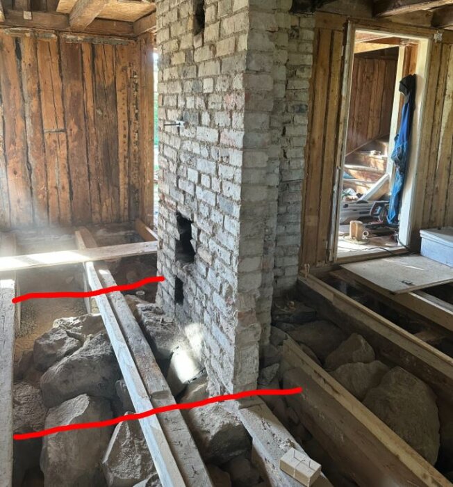 En äldre träramkonstruktion med synliga balkar och en murad skorsten, omgiven av stenar och byggmaterial i ett pågående renoveringsprojekt.