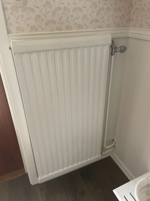 Vit radiator monterad på en vägg med blommig tapet, omgiven av vita lister och ett mörkt golv.