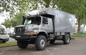 Grått terrängfordon med stora däck och ett rymligt lastutrymme, parkerat på en asfalterad väg med träd i bakgrunden.